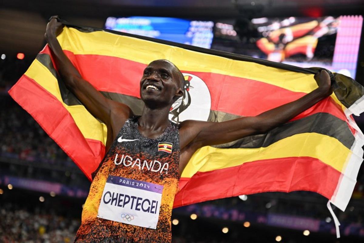 Winner Uganda's Joshua Cheptegei celebrates with the Uganda's national falg after crossing the finish line in the men's 10000m final of the athletics event at the Paris 2024 Olympic Games at Stade de France in Saint-Denis, north of Paris, on August 2, 2024. Uganda's Joshua Cheptegei sets a new Olympic record in the men's 10000m. Kirill KUDRYAVTSEV / AFP