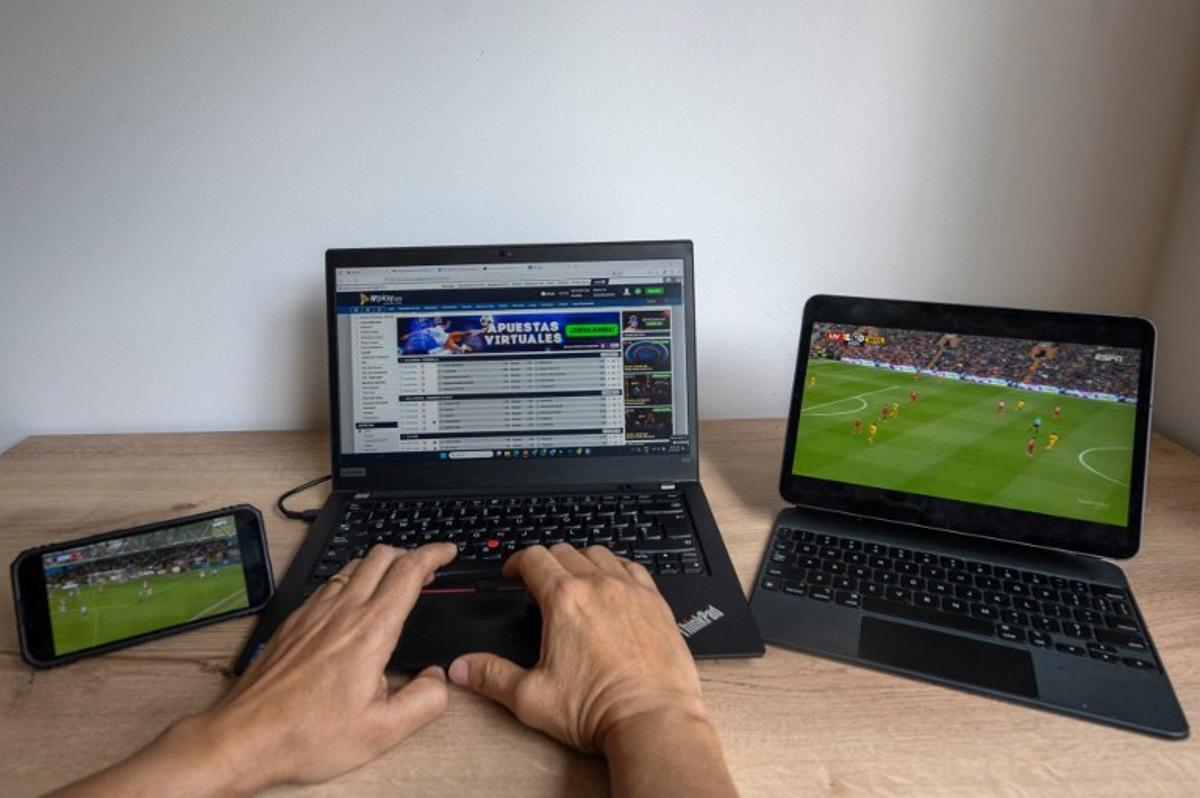 In this photo illustration taken on February 20, 2025, a user checks the betting company Wplay on a laptop while watching a football match in Bogota on February 17, 2025.  For the first time, online sports betting will pay sales tax in Colombia. The worst armed conflict crisis in decades prompted the government to tax this important sector of the economy in a decision that strikes a chord with football fans. Luis ACOSTA / AFP