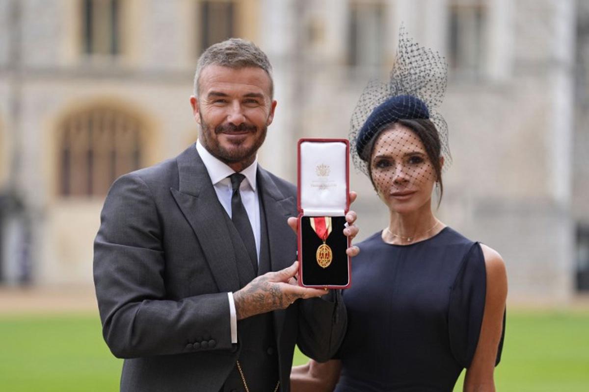 Former England footballer David Beckham (L) poses next to his wife singer and fashion designer Victoria Beckham (R) with his medal after being appointed as a Knight Bachelor (Knighthood) for services to sport and charity at an investiture ceremony at Windsor Castle on November 4, 2025.   Andrew Matthews / POOL / AFP